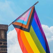 A square photo of the progressive pride flag flying near a chimney.